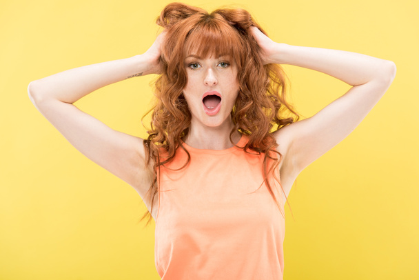 фронтальный вид потрясенной кудрявой рыжей женщины касающейся волос и смотрящей на камеру, изолированную на желтом
 - Фото, изображение