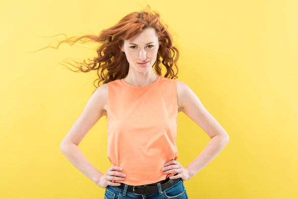 front view of attractive redhead curly woman with hands on hips looking at camera isolated on yellow - Φωτογραφία, εικόνα