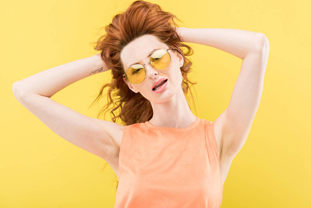 ενθουσιασμένος κοκκινομάλλα κορίτσι στα γυαλιά ηλίου αγγίζοντας σγουρά μαλλιά απομονωμένη στο κίτρινο - Φωτογραφία, εικόνα