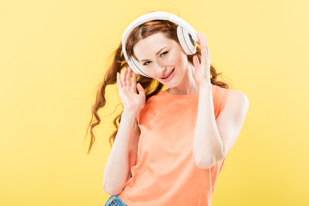 atractiva pelirroja escuchando música en auriculares y sonriendo aislada en amarillo
 - Foto, Imagen