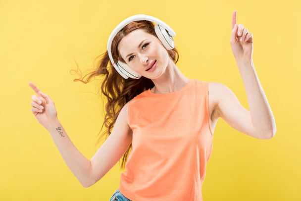 приваблива усміхнена руда жінка слухає музику в навушниках і танцює ізольовано на жовтому
 - Фото, зображення