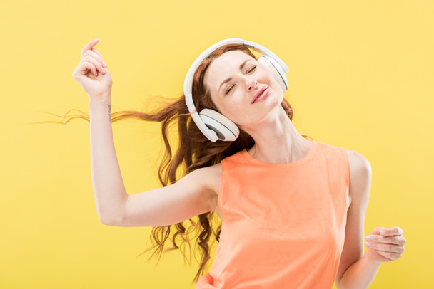 vonzó mosolygó vöröshajú nő hallgat zenét fejhallgató és táncoló csukott szemmel izolált sárga - Fotó, kép