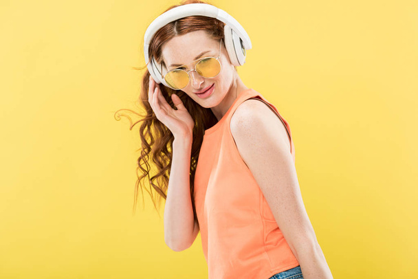 attractive redhead woman in sunglasses listening music in headphones and smiling isolated on yellow - Foto, Imagen