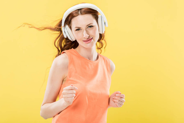 Vooraanzicht van aantrekkelijke redhead vrouw luisteren muziek in koptelefoon en glimlachend geïsoleerd op geel - Foto, afbeelding