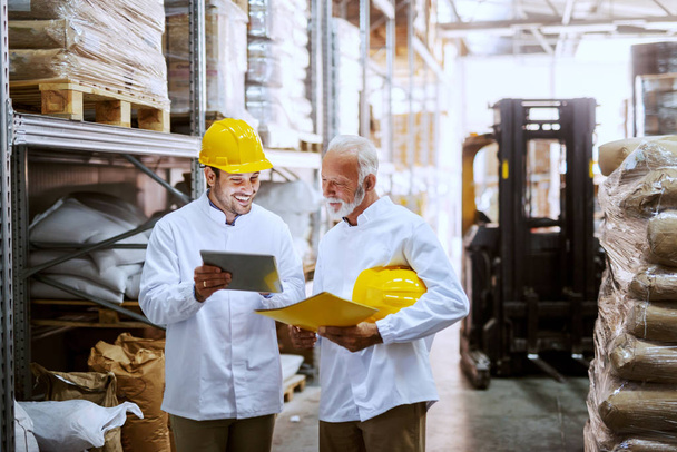 Smiling employees in white uniforms and yellow helmets on heads standing in warehouse and comparing data from tablet. Senior worker holding tablet while younger one holding yellow folder. - Photo, Image