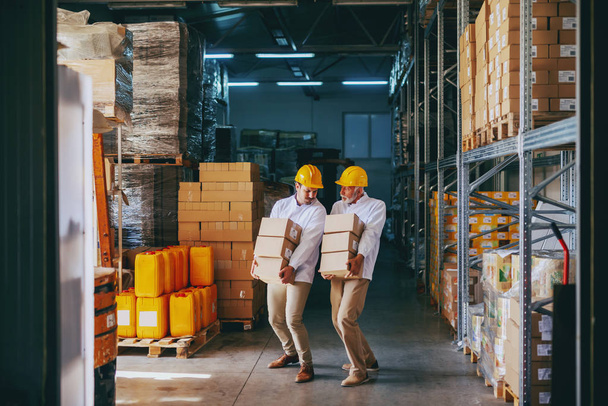 Dos trabajadores de cuello azul en uniformes blancos y con cascos amarillos en cabezas reubicando cajas pesadas en el almacén
. - Foto, imagen
