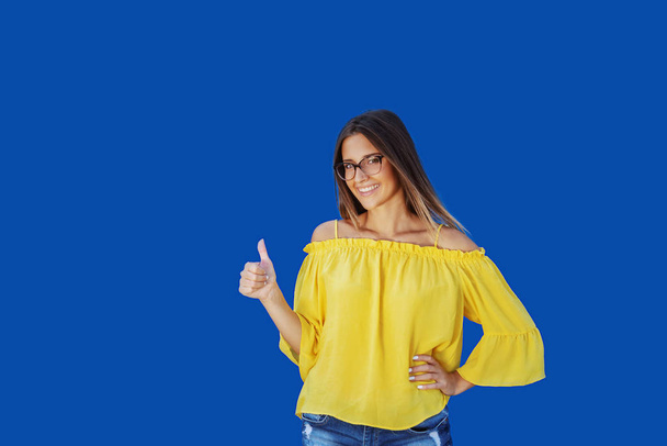 Souriant belle adolescente caucasienne en chemisier jaune, short en denim et avec des lunettes donnant pouces devant fond bleu
. - Photo, image