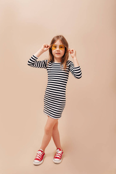 Full-lenght photo of happy little girl wearing stripped dress and round stylish glasses has fun over isolated beige background - Photo, Image
