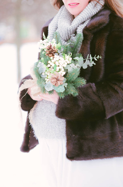 A young pregnant woman in a mink coat and a long dress with a bouquet in winter on the snow in the park - Foto, imagen