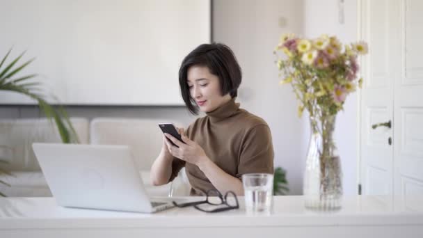 Азіатські жінки прокручування телефону сидять у білому домашньому офісі - Кадри, відео