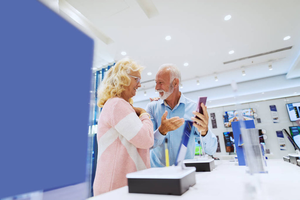 Cute smiling senior couple standing in tech store and choosing new smart phone. - Photo, Image