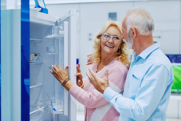 Cute happy smiling Caucasian senior choosing new refrigerator for their home. Tech store interior. - 写真・画像
