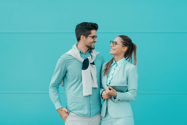 Cheerful multicultural couple dressed elegant looking at each other and standing in front of turquoise background. Woman holding tablet. - Photo, Image