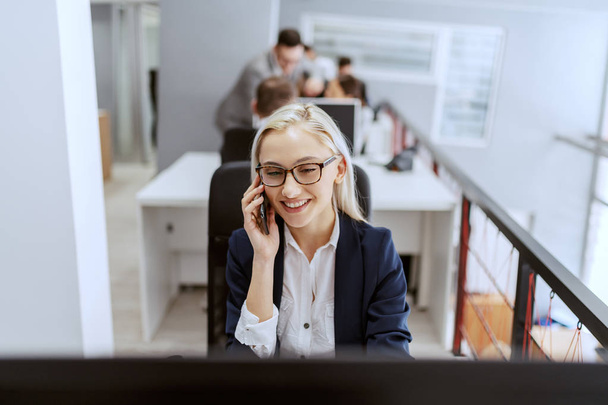 Portrait of smiling Caucasian blonde female employee dressed in formal wear and with eyeglasses using computer while taking on the phone. - Φωτογραφία, εικόνα