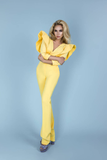 Elegant blonde woman in stylish yellow jumpsuit and fashionable accessories on color background. Beautiful fashion model on blue background. - Photo, Image