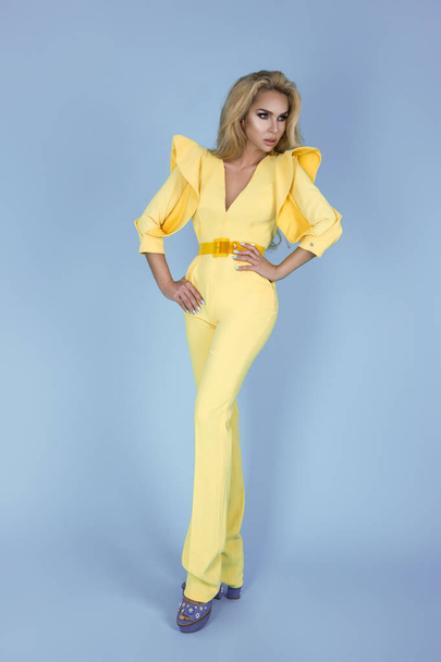 Elegáns szőke nő stílusos sárga JumpSuit és divatos kiegészítők színes háttér. Gyönyörű divatmodell kék háttérrel. - Fotó, kép