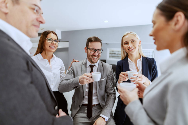 Bir grup gülümseyen neşeli iş arkadaşları, işyerinde otururken ve dururken, duraksarken sohbet edip kahve içerken. - Fotoğraf, Görsel