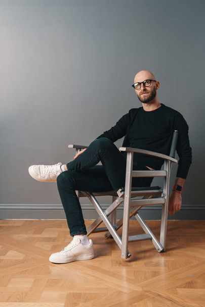 Um homem moderno sentado em uma pose confiante, perna sobre perna em uma cadeira, em fundo de parede cinza no estúdio
 - Foto, Imagem