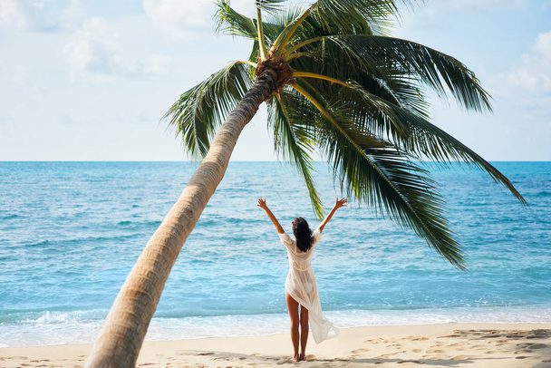 Pohled na šťastnou mladou ženu si vychutnejte dovolenou na tropické pláži, stojící pod palmovým stromem. Letní koncept   - Fotografie, Obrázek