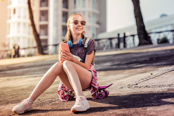 Smiling little lady wearing sunglasses and headphones while staying outside - Foto, afbeelding