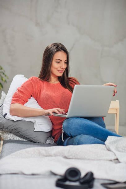 Gyönyörű kaukázusi barna ül az ágyon a hálószobában, és használ laptop. - Fotó, kép
