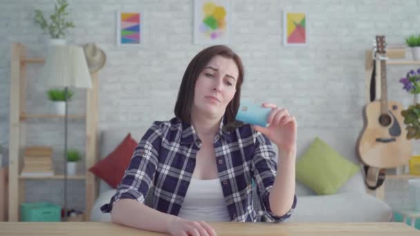 Sad young woman with credit card in hand sitting at a table in a modern apartment,concept the idea of lack of money - Footage, Video
