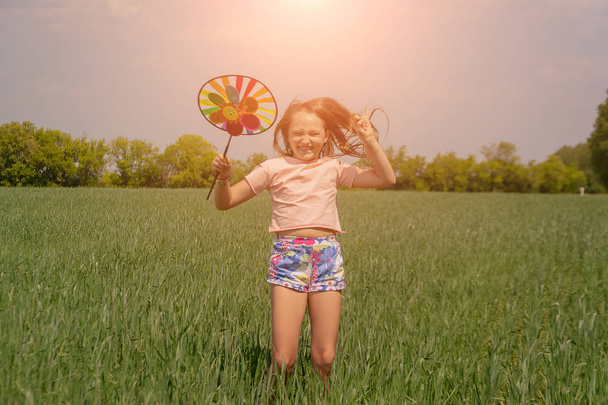 Szczęśliwy dziewczyna z długimi włosami trzyma kolorowy wiatrak zabawka w jej ręce i skoki. - Zdjęcie, obraz