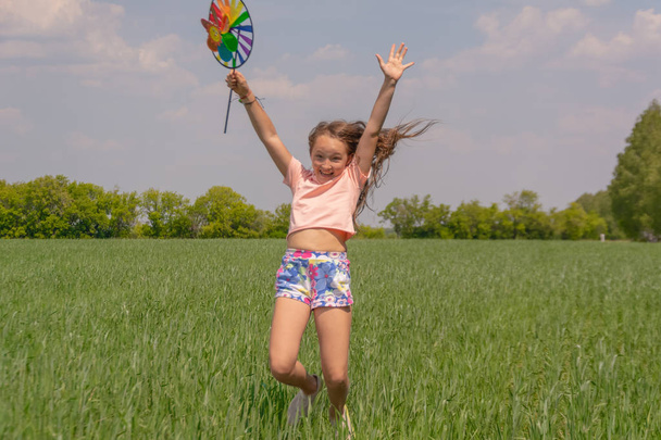 Happy girl with long hair holding a colored windmill toy in her hands raises her hand and jumps. - Photo, Image
