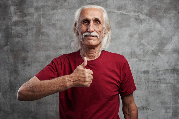 Modern gray-haired caucasian old man with a mustache in red t-shirt shows thumb up and smiles, joyful happy face against gray wall - Φωτογραφία, εικόνα