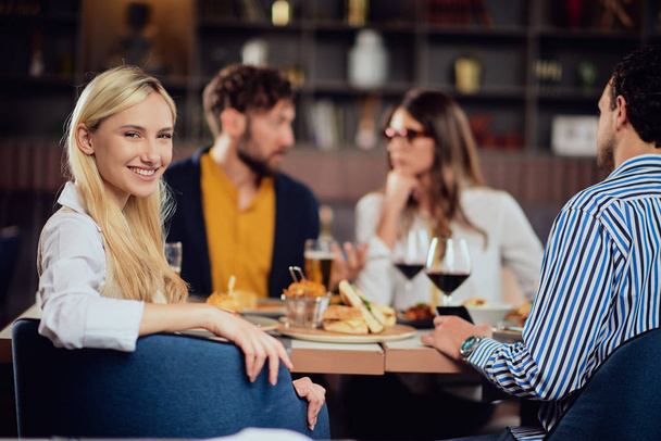 Jeune femme mignonne blonde caucasienne souriante habillée smart casual assis à la table dans le restaurant et regardant par-dessus l'épaule. En arrière-plan sont ses amis qui dînent
. - Photo, image