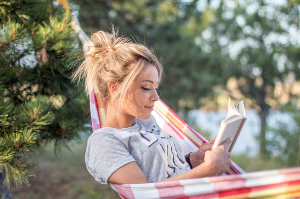 young attractive caucasian woman reading book in hammock in the forest, lake on the background - 写真・画像
