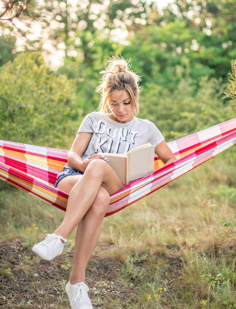 young attractive caucasian woman reading book in hammock in the forest, sun on the background - Foto, imagen