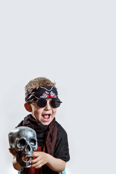 un niño rockero en gafas negras, bufanda, bandana y con un cráneo en las manos sobre un fondo claro
 - Foto, Imagen