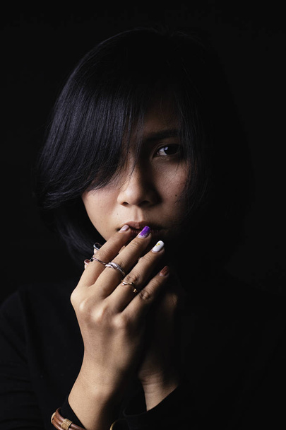Aasialainen lyhyet hiukset tyttö yllään musta pitkähihainen paita ja koruja valmistettu metallista ja nahasta, surullinen kasvot musta tausta ja varjot
 - Valokuva, kuva