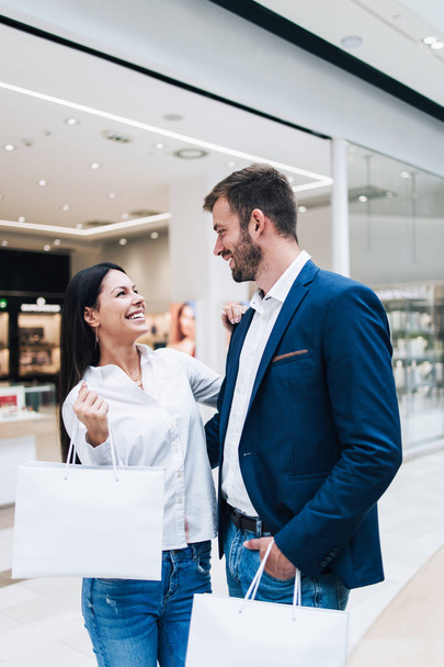Beautiful couple enjoying in shopping mall or center. They standing in front of huge modern store, smiling and looking at side while holding shopping bags. Shoot from below. - Фото, изображение