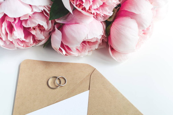 Обручальные кольца, конверт, цветы пионов на белом фоне, ко
 - Фото, изображение