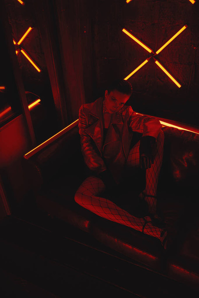 cyberpunk shooting of model wearing red bikers jacket sitting in leather sofa against neon - 写真・画像