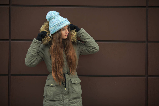 Portrait of a young pretty caucasian teenage girl in a blue hat, black gloves and a green jacket with a hood in the winter outside. - Фото, изображение