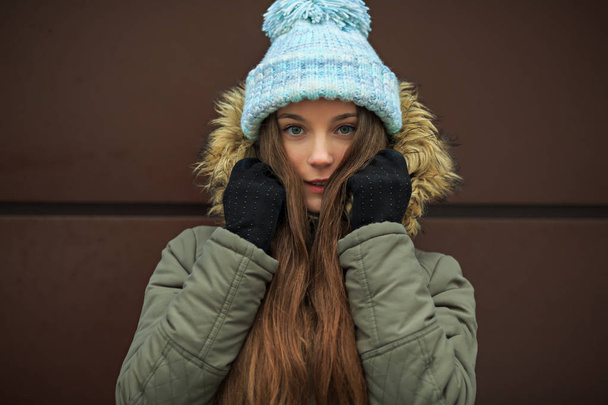 Portrait of a young pretty caucasian teenage girl in a blue hat, black gloves and a green jacket with a hood in the winter outside. Closeup. - Fotó, kép