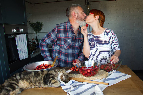 大人のカップルの男女はイチゴジャムのためにイチゴの皮をむき、お互いを養い、笑い、楽しみを持って、メインクーン子猫は台所のテーブルの上にあります - 写真・画像