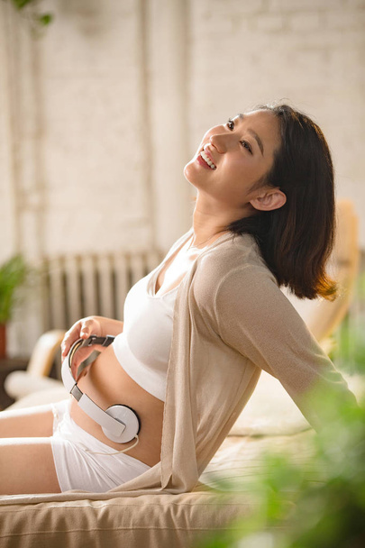 boldog fiatal terhes nő, aki a fejhallgatót a hasa, és nevetnek otthon - Fotó, kép