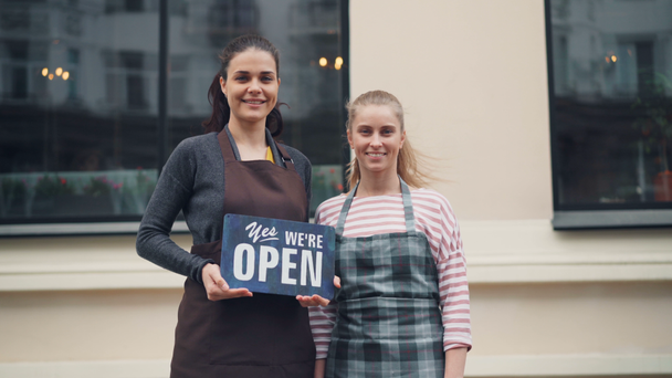 Portrait of pretty young women holding open sign standing outdoors near new cafe - Footage, Video