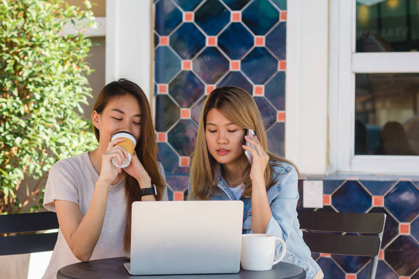 Mujeres jóvenes asiáticas que trabajan en el ordenador portátil usando y mirando teléfono inteligente y beber café mientras están sentadas en la cafetería. Estilo de vida de las mujeres de comunicación y trabajo en concepto de cafetería
. - Foto, imagen