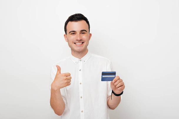 Wesoły młody człowiek pokazując kciuk w górę gest i z innej ręki posiadania karty kredytowej na białym tle - Zdjęcie, obraz