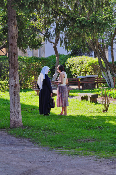 Kaksi naista puhuu puun varjossa aurinkoisena päivänä. Mitä? Toinen on nunna kirkkopuvuissa, toinen on seurakuntalainen.
. - Valokuva, kuva