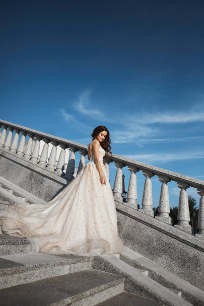 Beautiful, sexy and fashionable brunette girl in white lace wedding dress posing on the stairs with blue summer sky at background - Photo, image