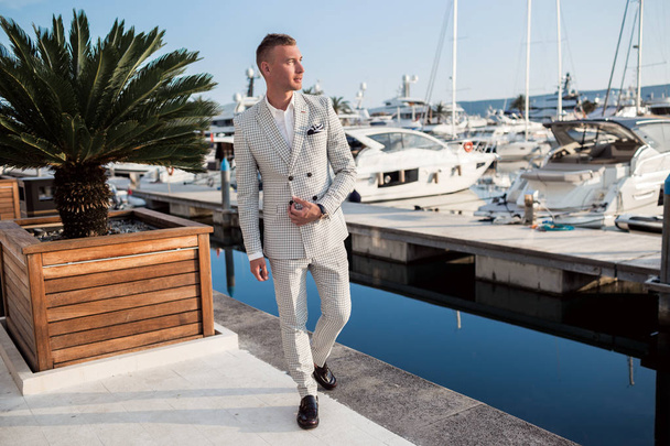 Stylish businessman is wearing white fashion costume stands on the background of the bay with luxury yachts. Montenegro bay. - Zdjęcie, obraz