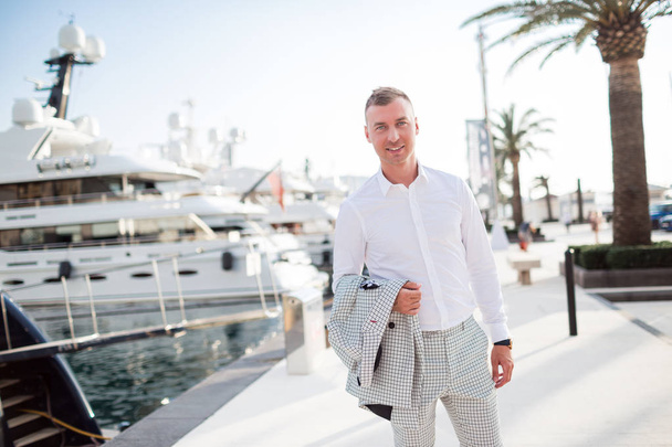 Stylish businessman is wearing white fashion costume stands on the background of the bay with luxury yachts. Montenegro bay. - Foto, Imagem