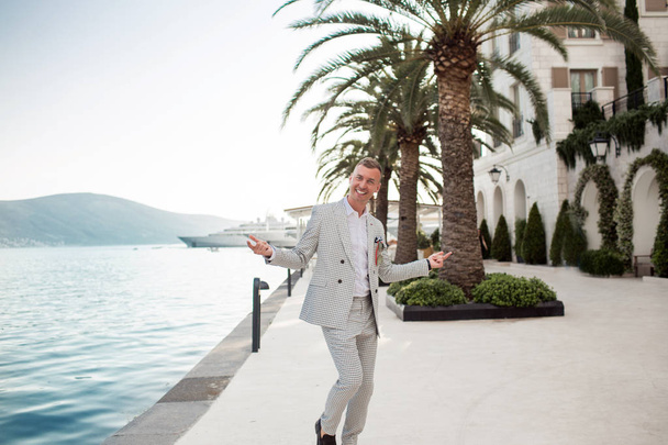 Stylish businessman is wearing white fashion costume stands on the background of the bay with luxury yachts. Montenegro bay. - Fotó, kép