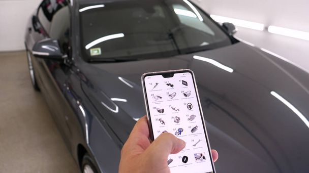 Against the background of a car in a garage, a man orders parts through a mobile device by selecting different parts on the Internet. Concept of: Smartphone, Order, Online Details, Engine, New. - Foto, Imagem